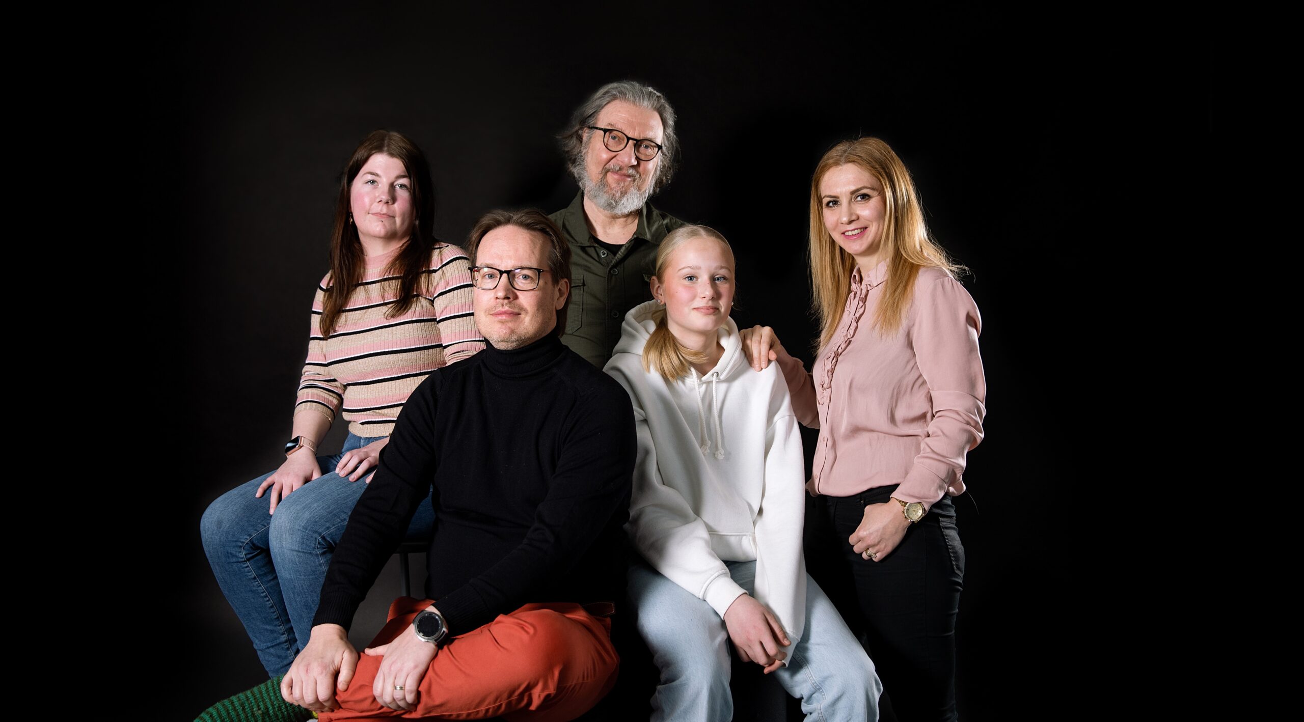 Viisi eri ikäistä hymyilevää henkilöä kuvattuna mustaa taustaa vasten. Kuvituskuva.